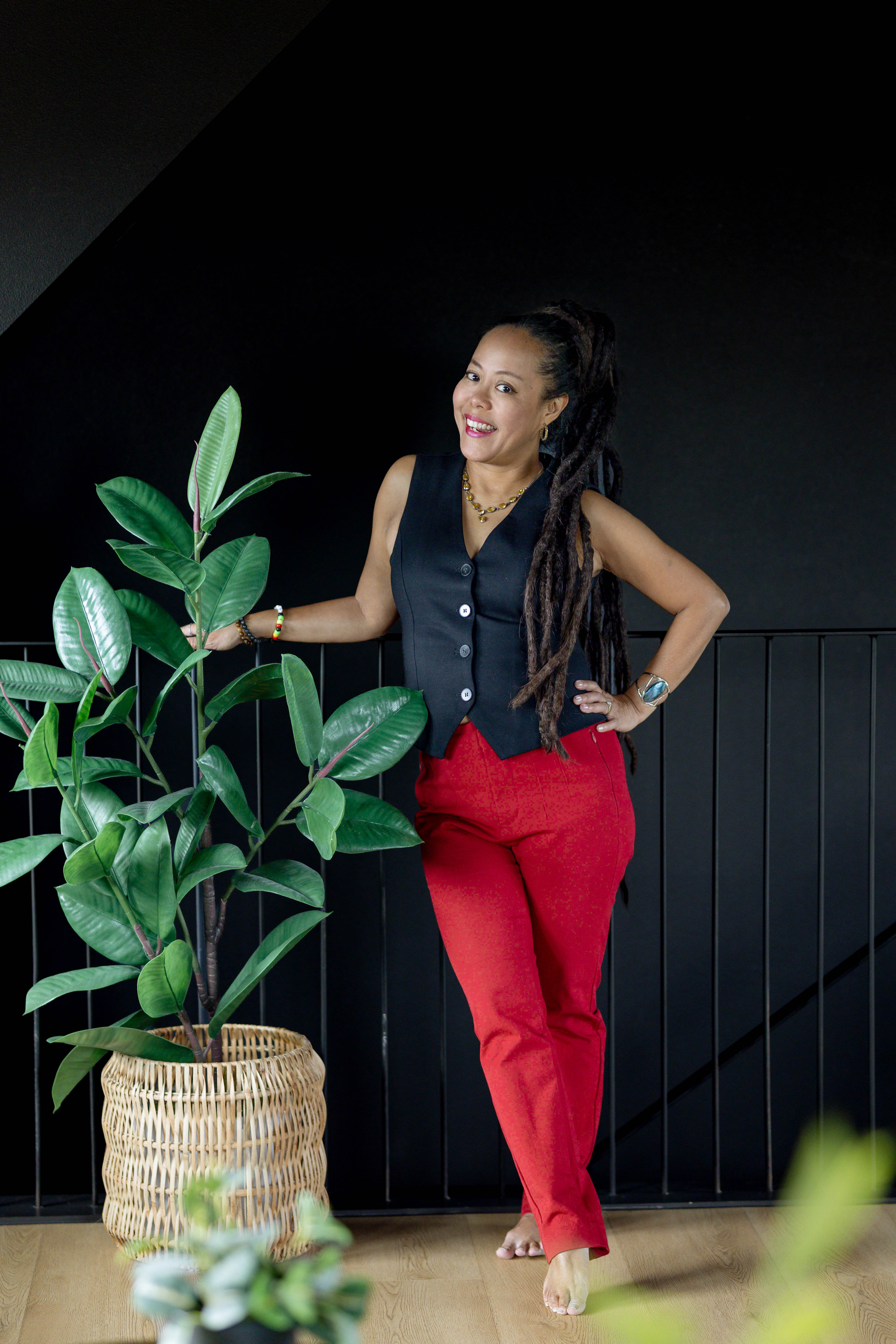 Portrait of Anne Marie Pizarro smiling beside a leafy plant against a dark background.