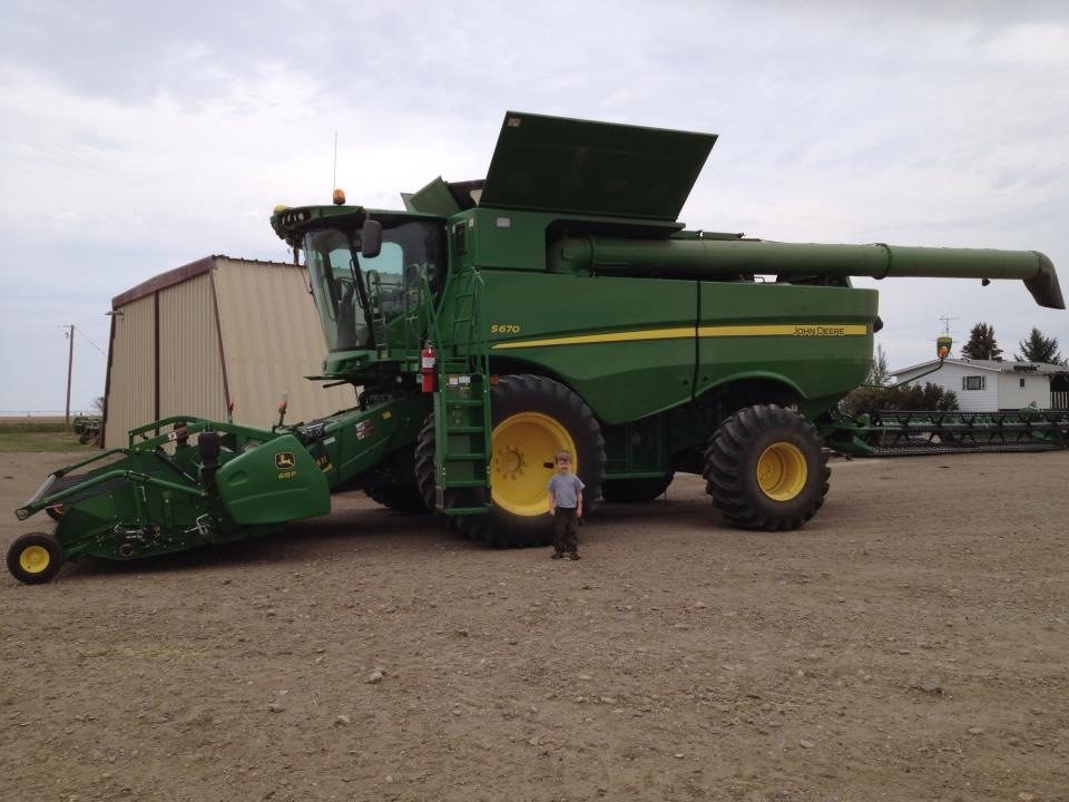 A John Deere S670 combine with a pickup header, with a young visitor beside the wheel.
