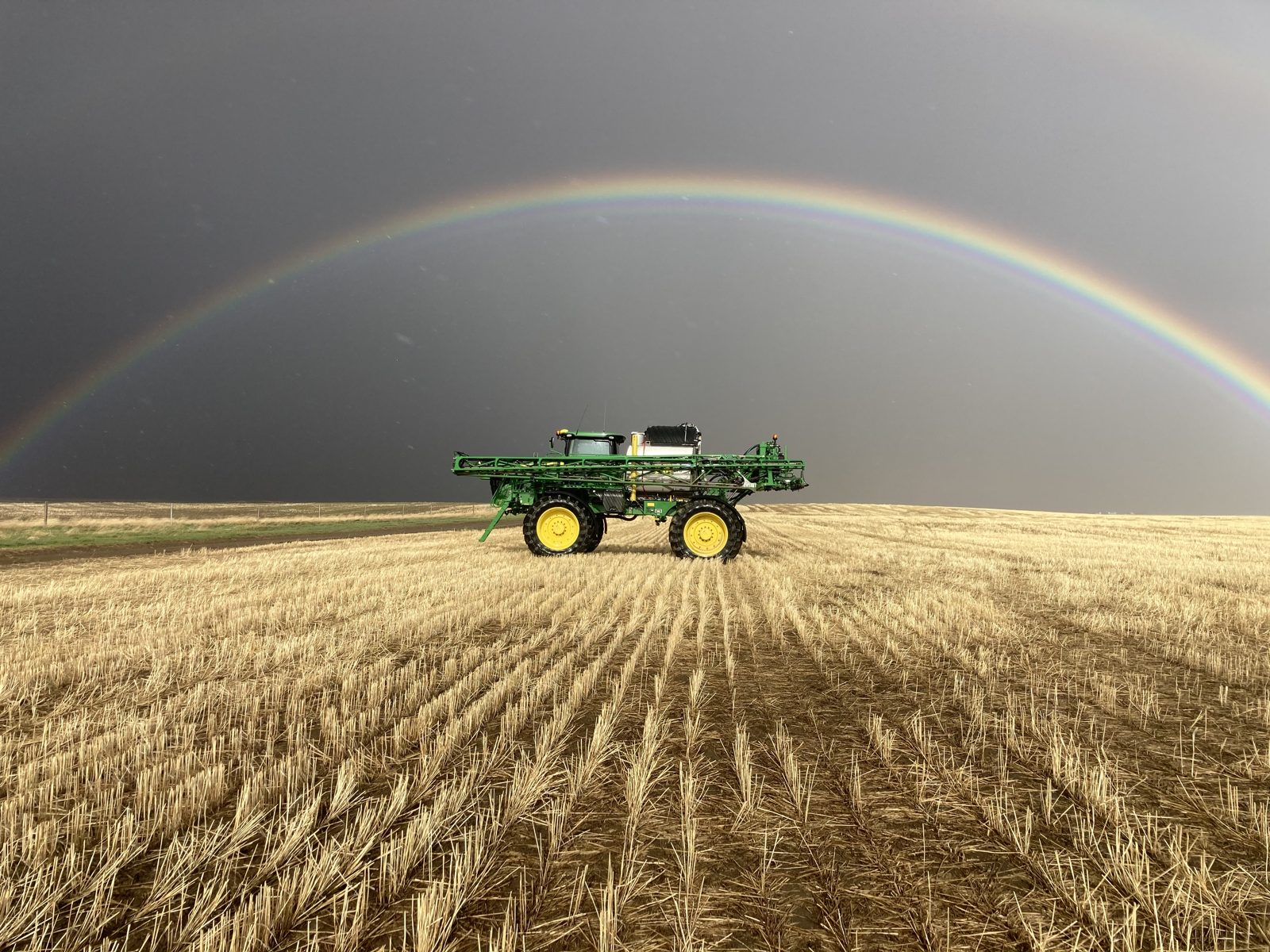 A John Deere crop sprayer beneath a full rainbow.