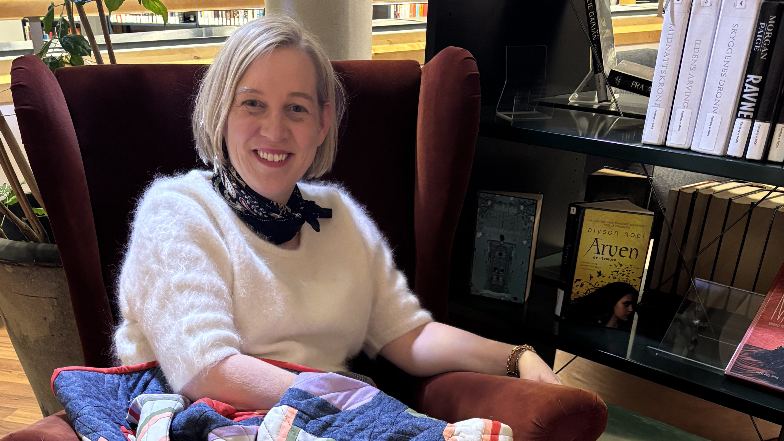 Solveig i en lenestol med en quilt på biblioteket