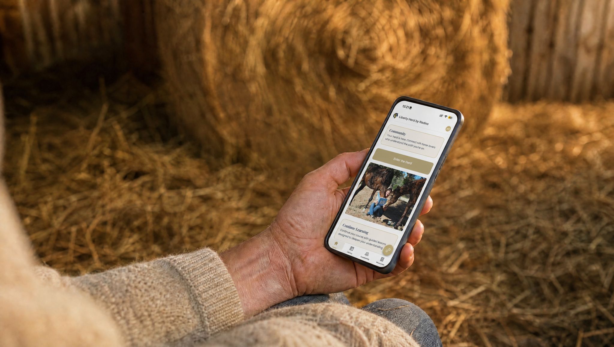 A weathered hand in a cream knit jumper holding a phone showing the Liberty Herd app, its Community panel and Enter the Herd button on screen, against sunlit hay bales in a barn