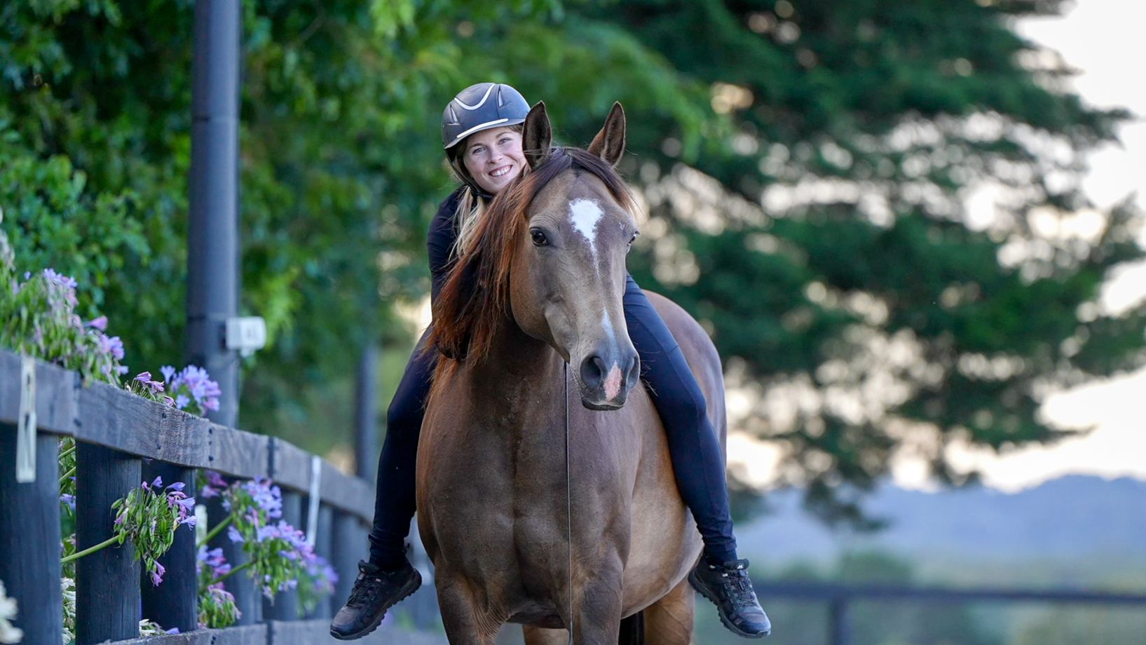 Nadine riding bareback and bridleless on a bay horse past a fence line of purple agapanthus, leaning forward and smiling