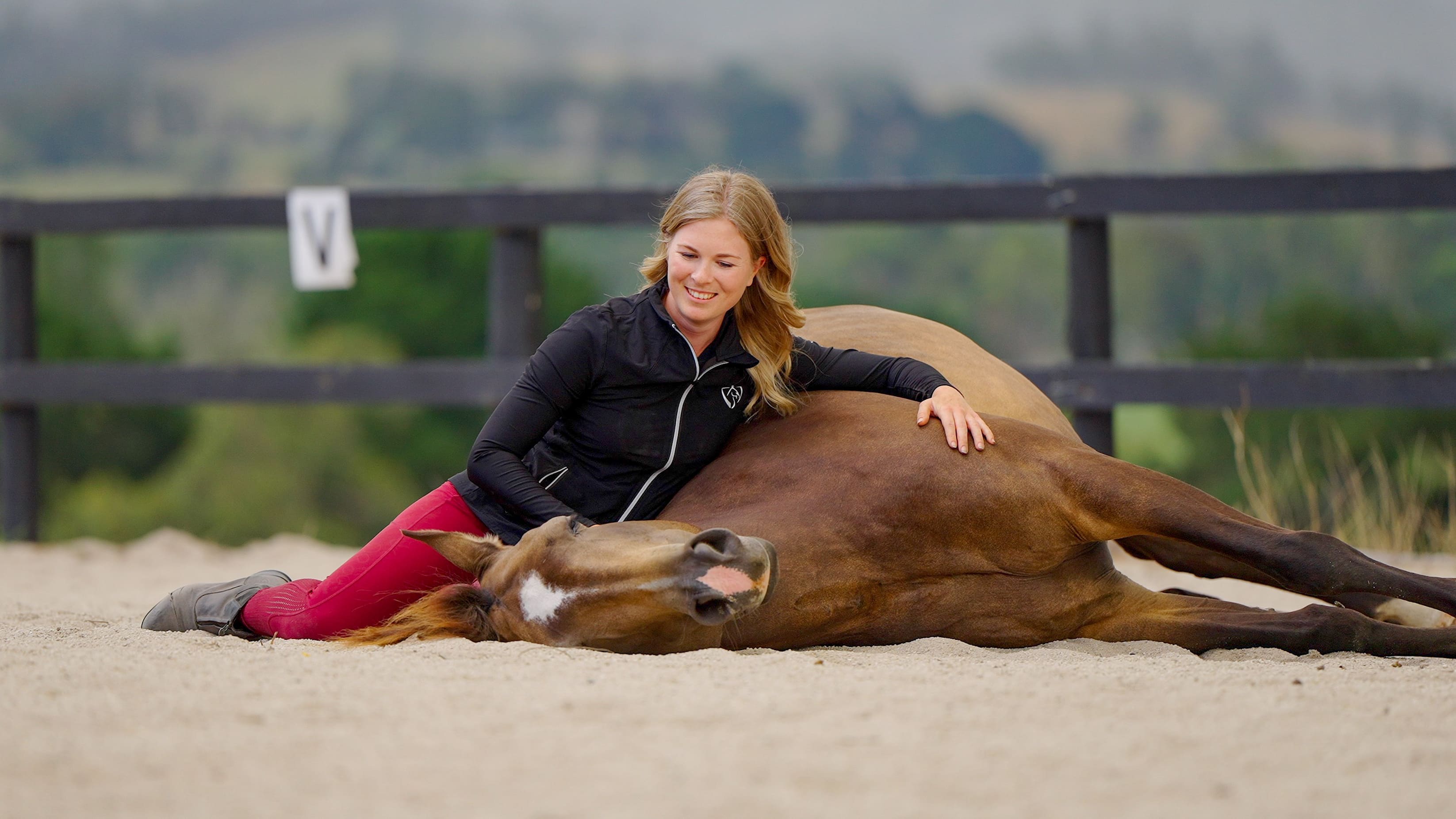 Nadine sitting in the sand beside her horse, who is lying flat out and completely relaxed