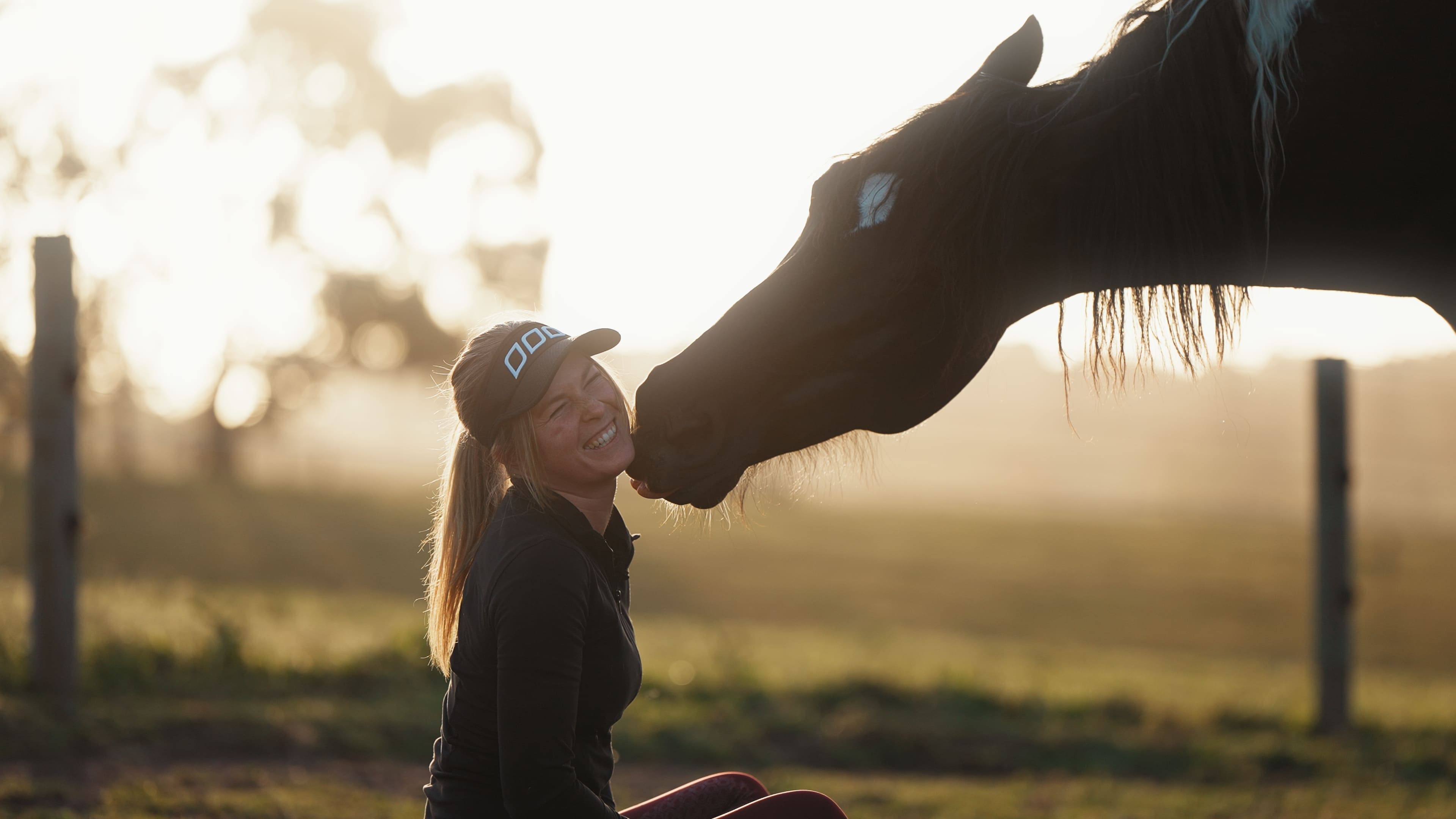 A woman kneeling in golden evening light, smiling, as her dark horse reaches down to touch her face