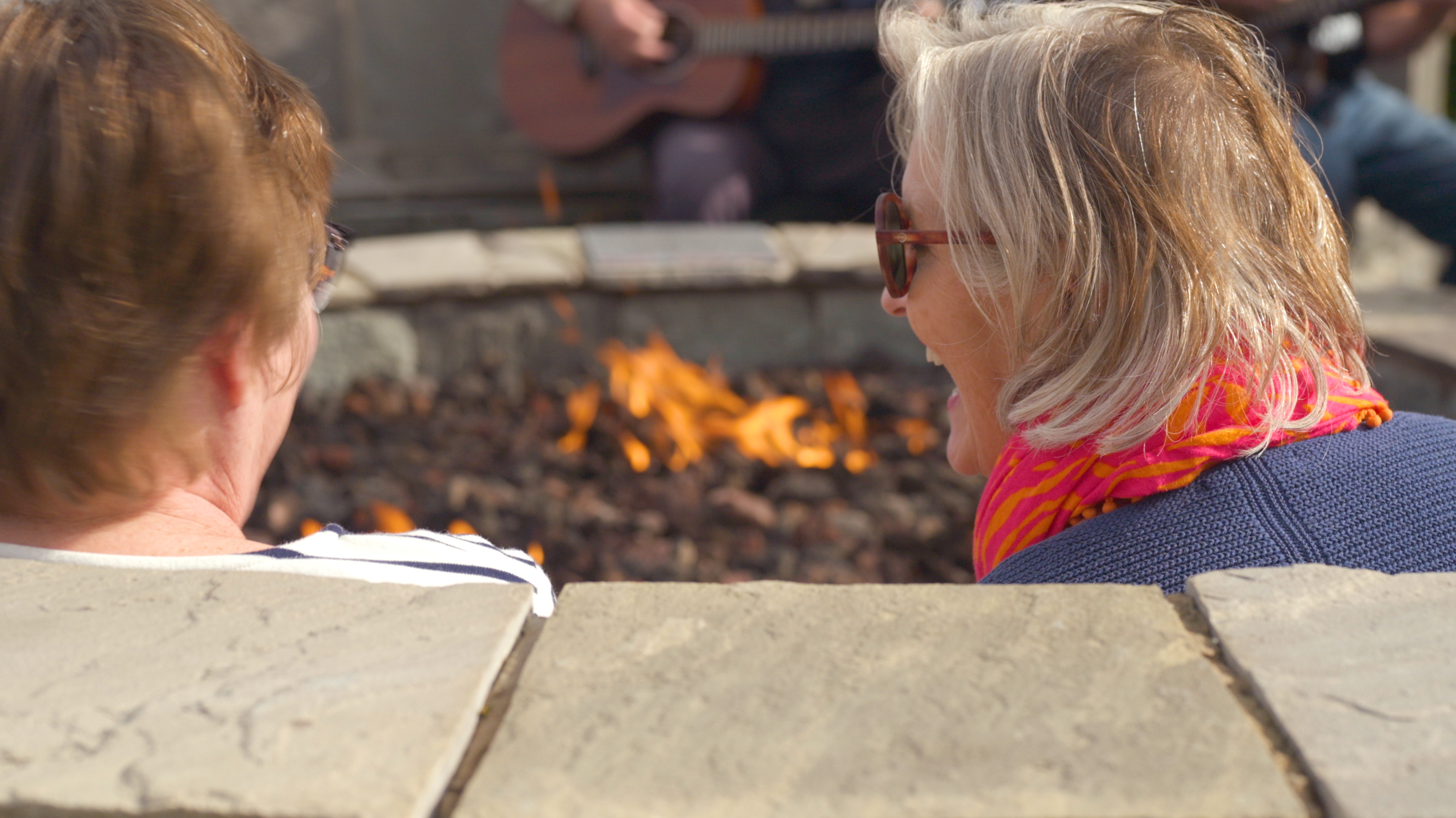 Two guests talking beside a fire pit while a guitar is played behind them