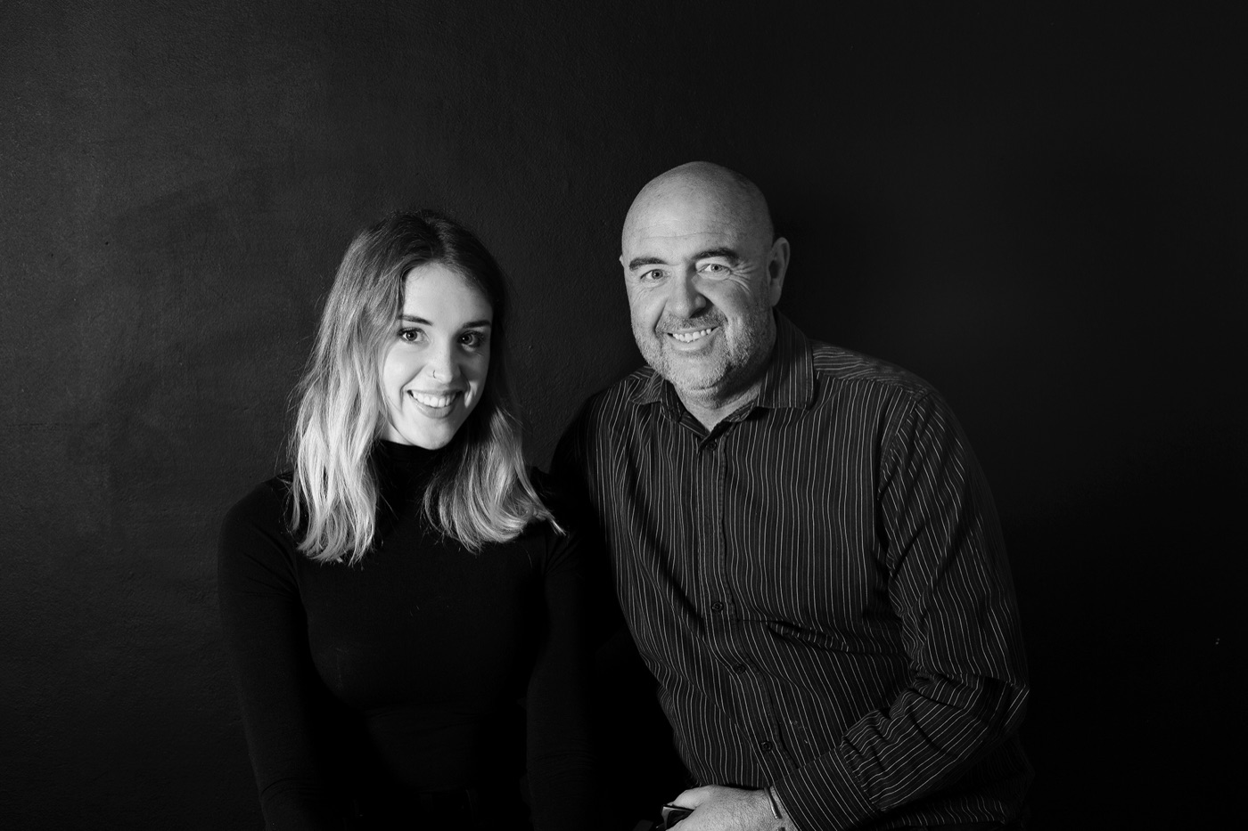 Black and white portrait of Samantha Lee Osner and Martin Osner standing together against a dark wall.