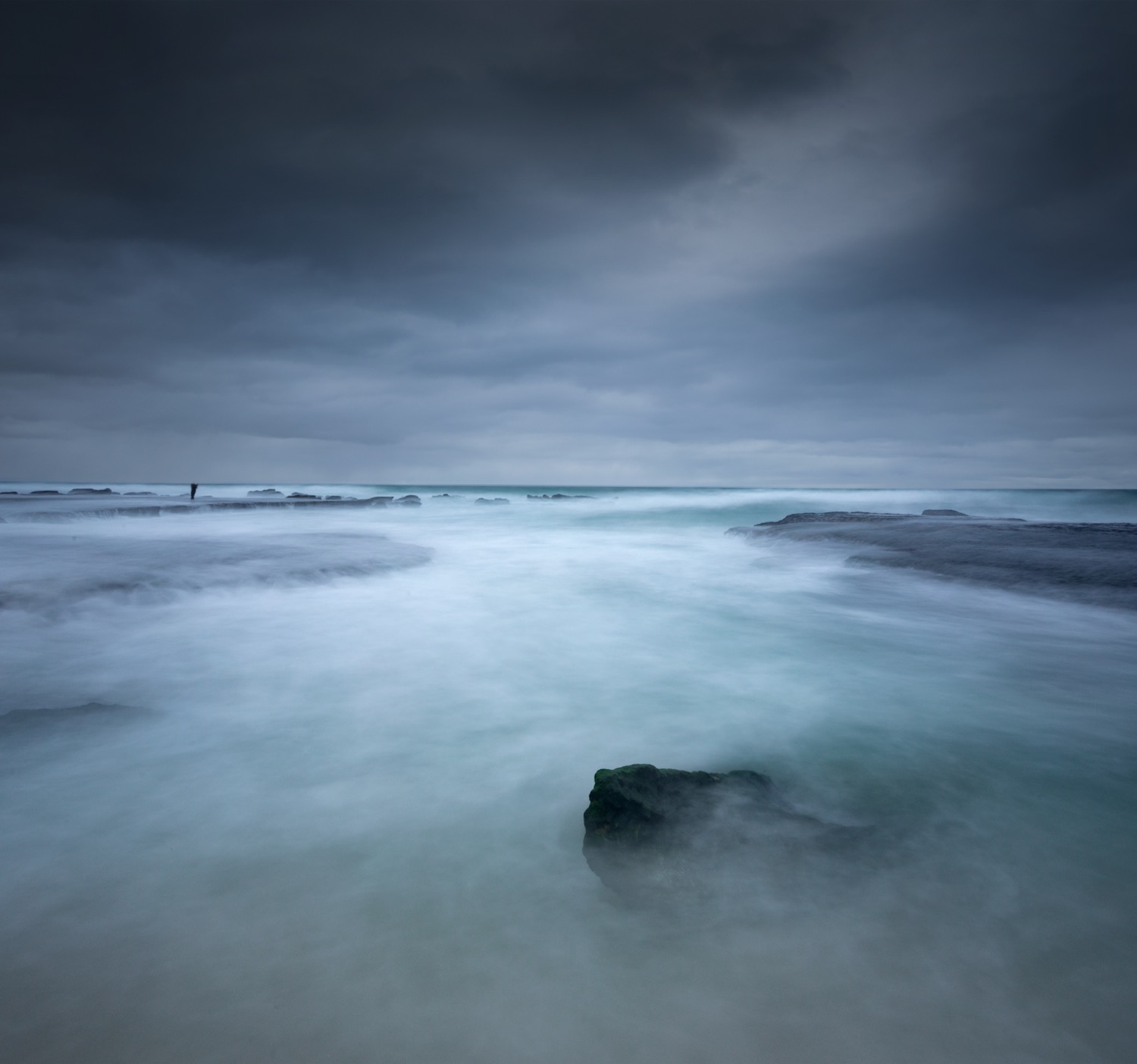Dark rock sitting in pale moving water under a heavy blue evening sky