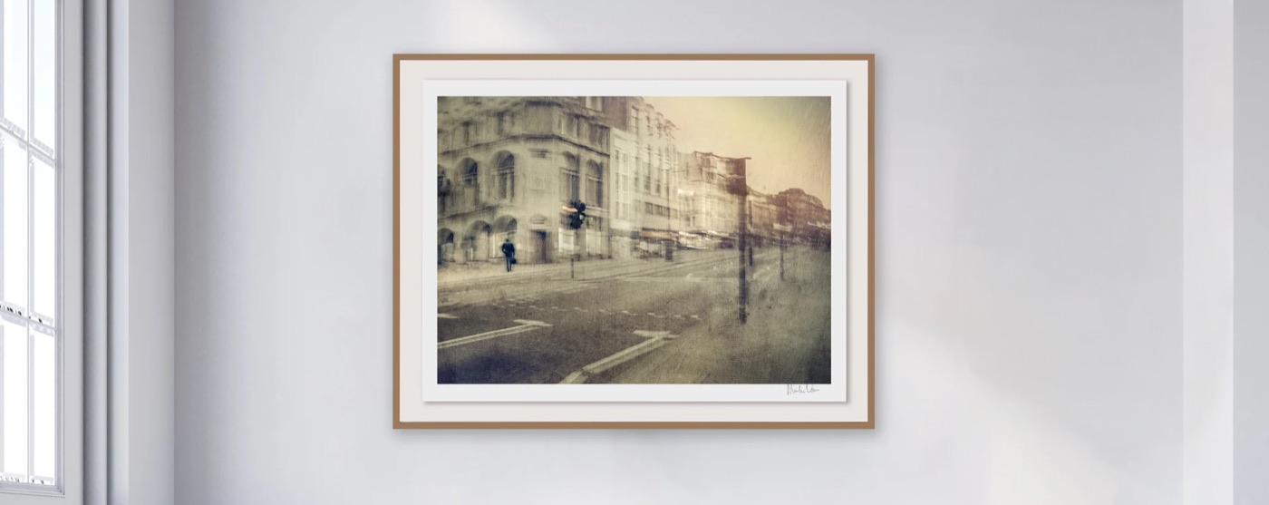 Démodé No.1 by Martin Osner, a framed fine art photograph hanging on a pale gallery wall, showing a blurred sepia toned street of old buildings with a single figure crossing the road.