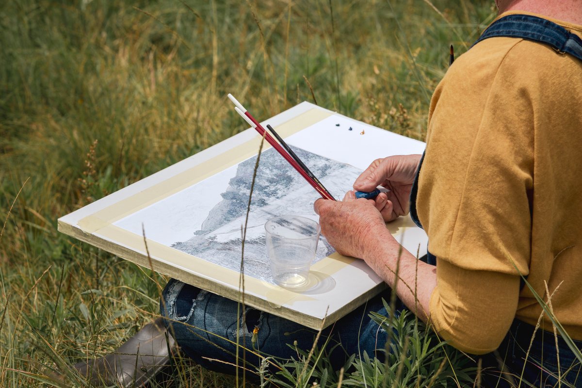 A student painting on a board in the field with mountains behind