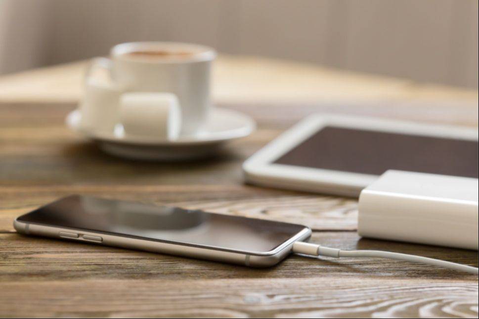 A phone charging on a wooden kitchen table, away from the bedroom, beside a cup of coffee and a tablet.