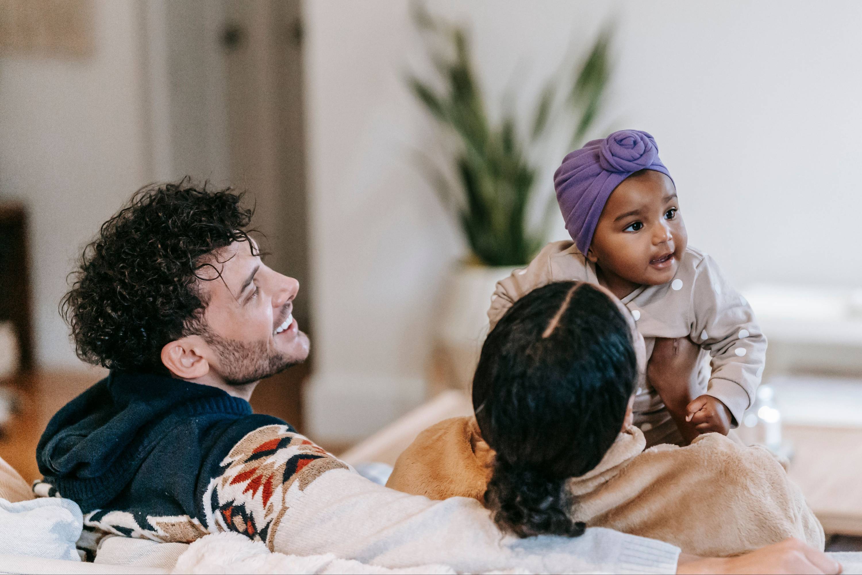 Two parents lying on a bed with their baby sitting up beside them, all three relaxed and unhurried.