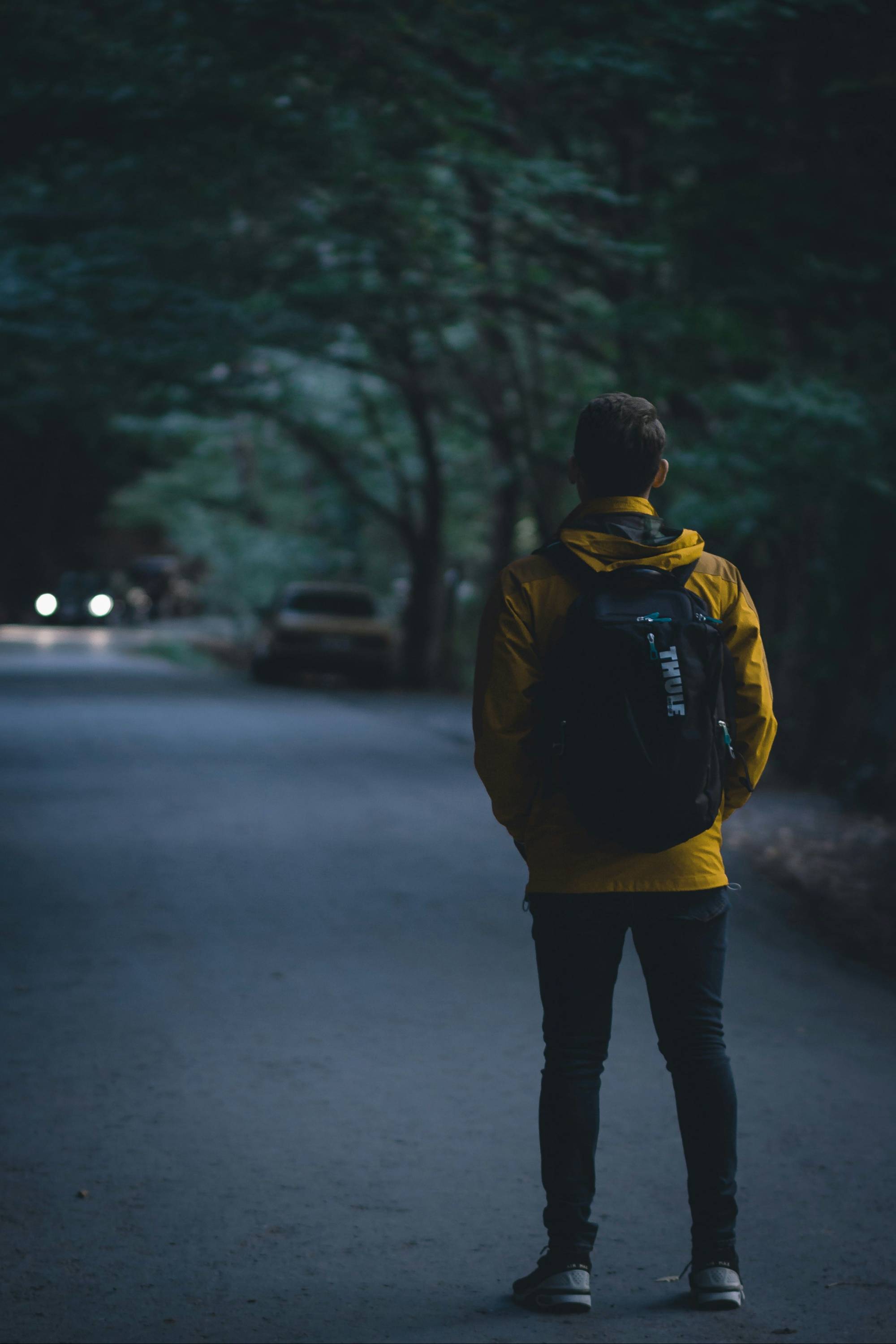 A teenager in a yellow jacket with a backpack, standing alone on a dark tree-lined road, with car headlights approaching in the distance.