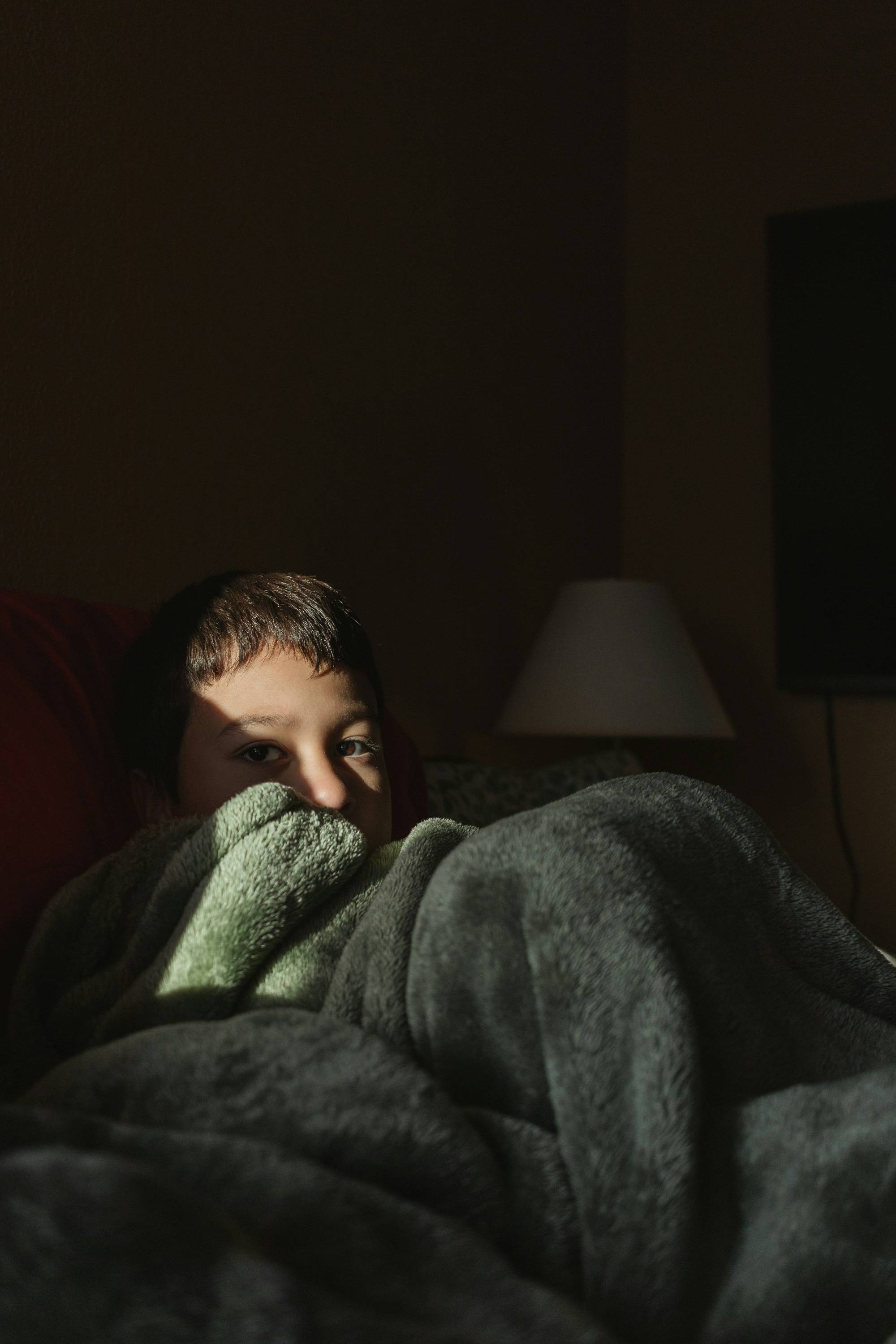 A school-age child lying awake in bed at night, covers pulled up, with a lamp lit in the dark room behind them.