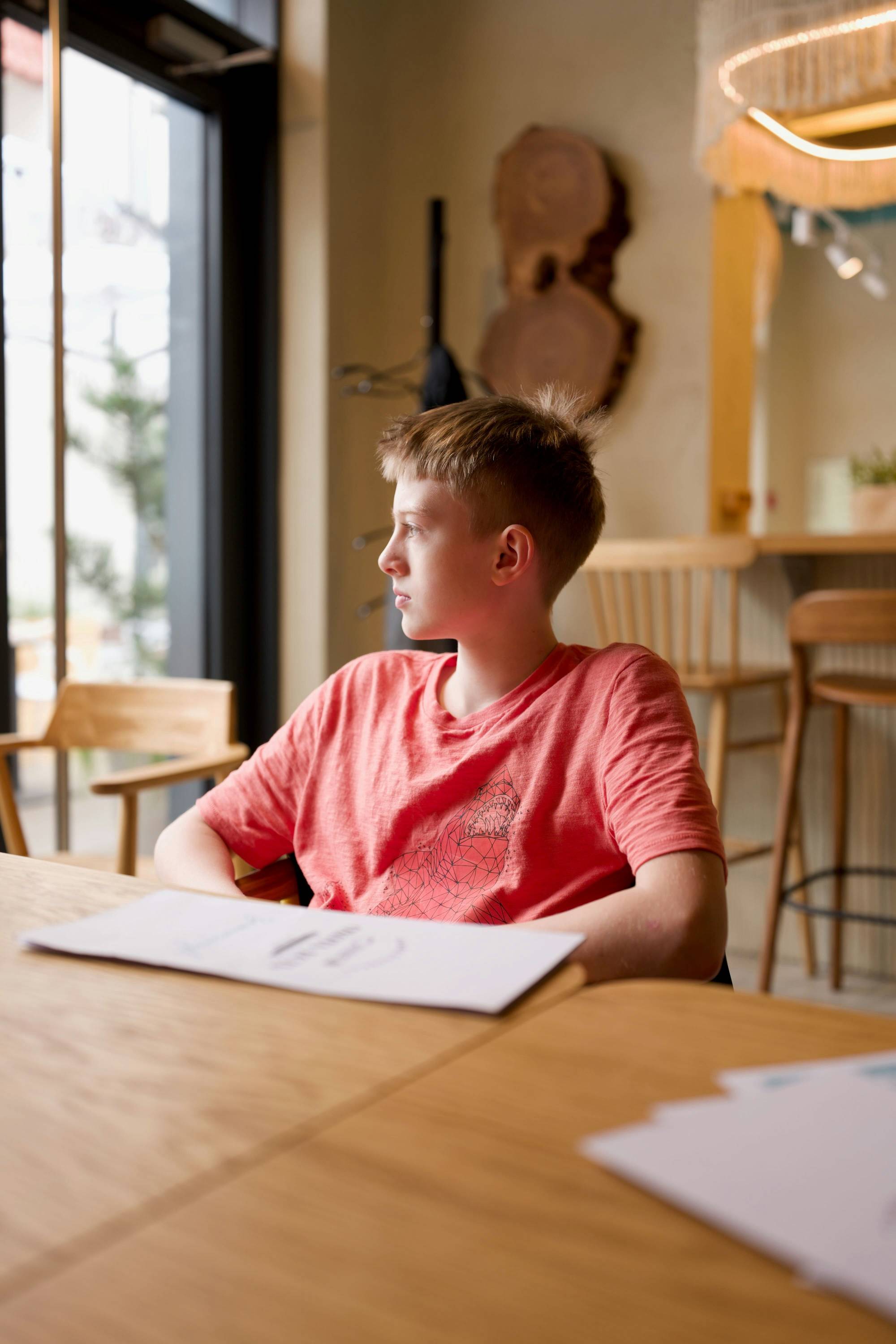 A school-age child sitting at a wooden table with papers in front of him, looking away, attention somewhere else.