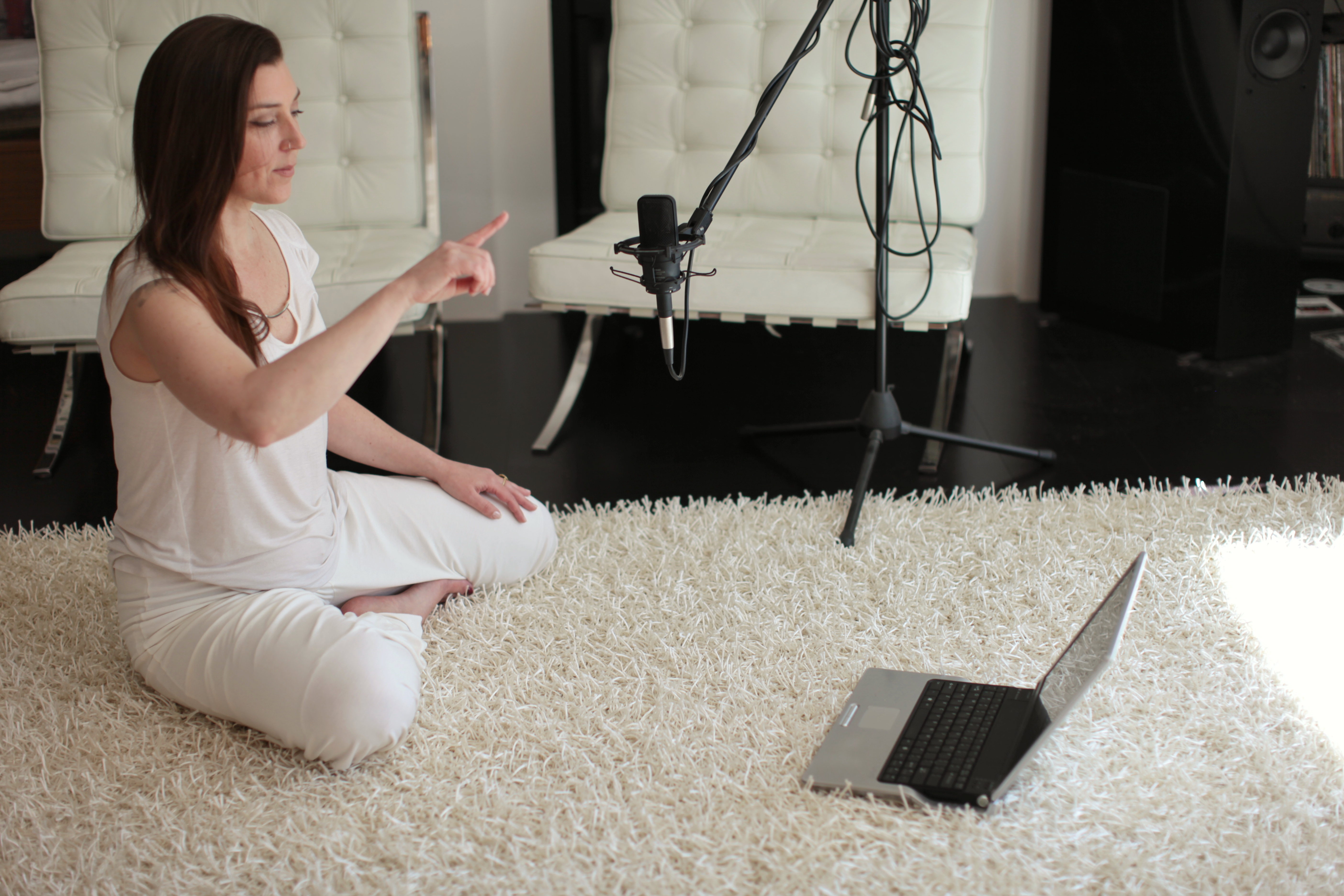 Marilyn seated on a soft rug, guiding a practice with one hand raised, a microphone and laptop set up beside her