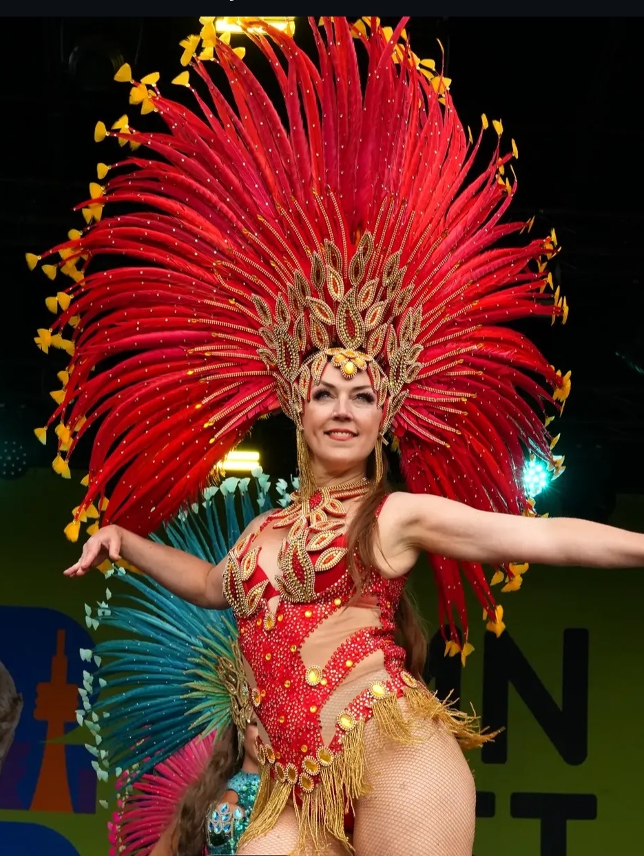 Marilyn performing Brazilian carnival dance on stage in a red feathered headdress, arms open wide