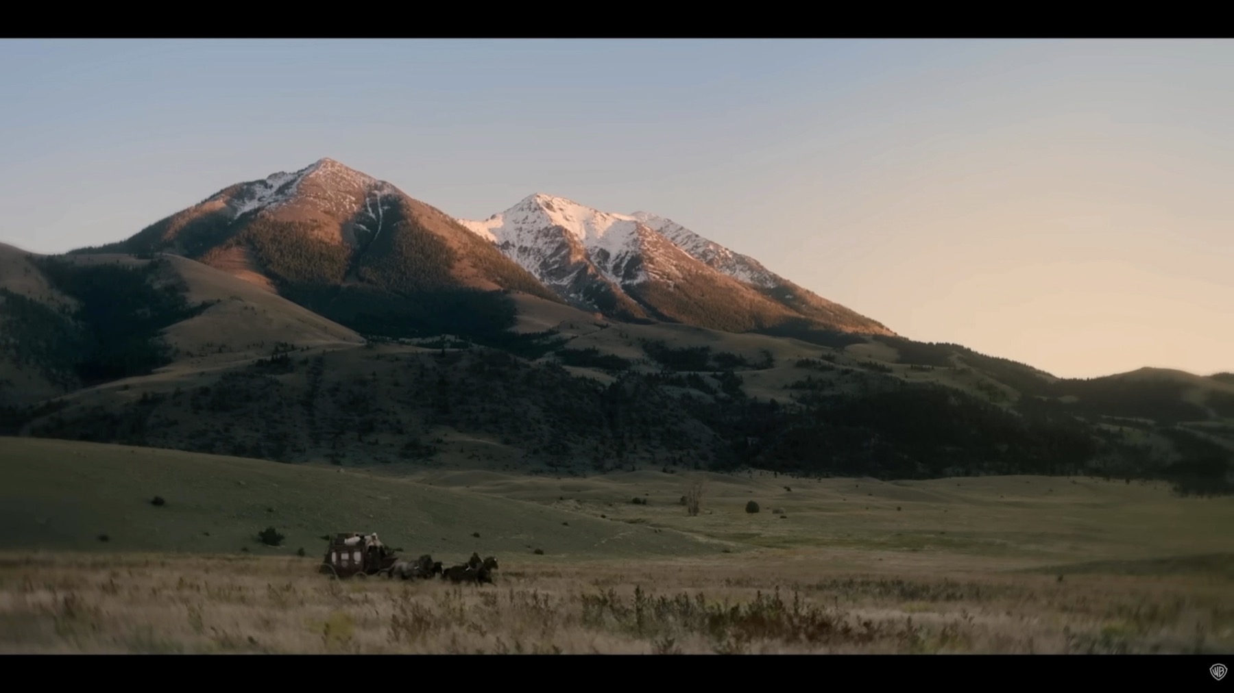 Wide landscape frame from The Unholy Trinity with mountains, open grassland, and a wagon placed low in the composition