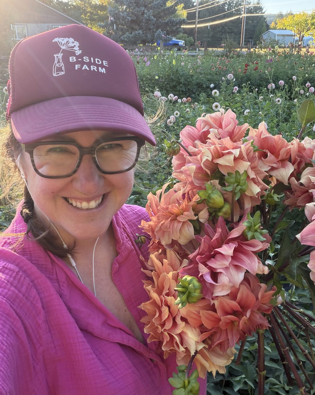 Lennie in a B-Side Farm cap holding an armful of peach dahlias, dahlia field behind her in evening light.