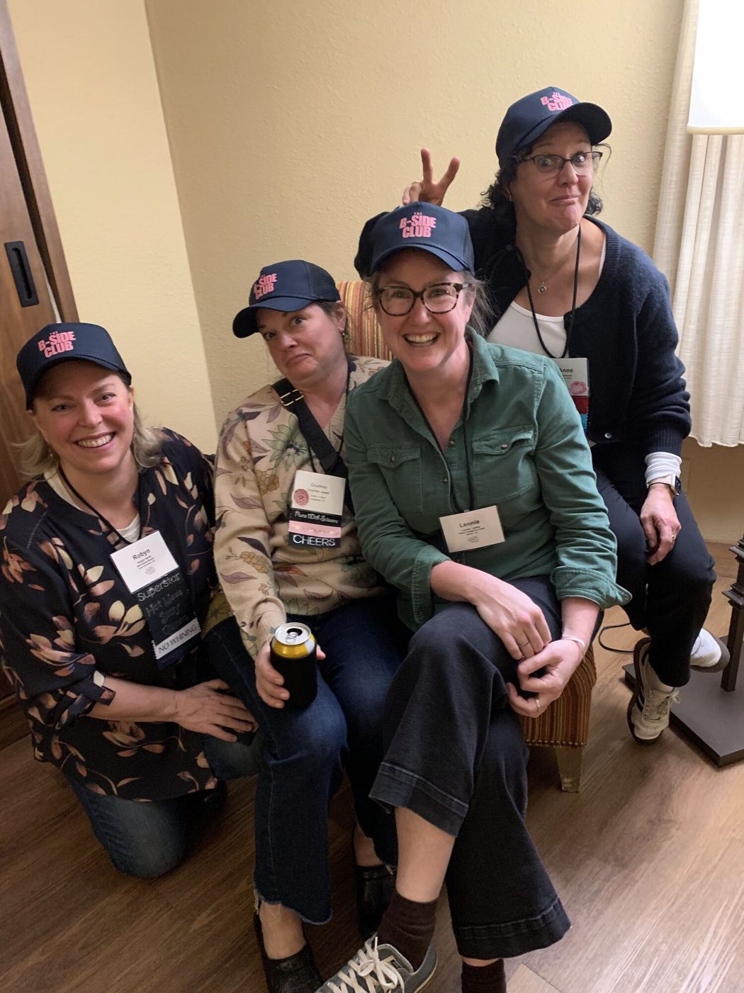 Four B-Side Club members in navy club caps and name badges, crowded together grinning, one holding up bunny ears behind another.