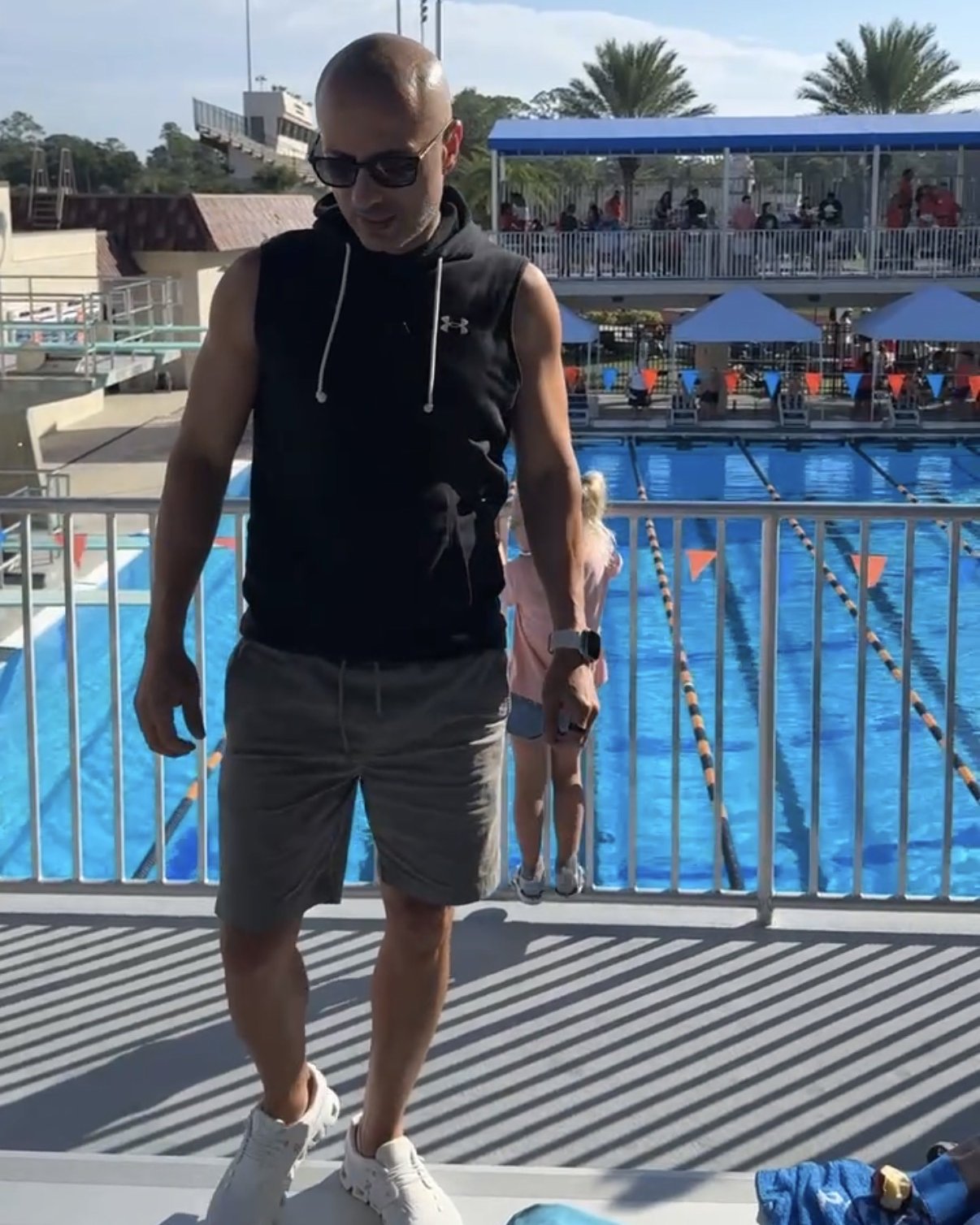 Feras in sunglasses and a black hoodie standing on the bleachers at an outdoor pool during his son's swim lesson