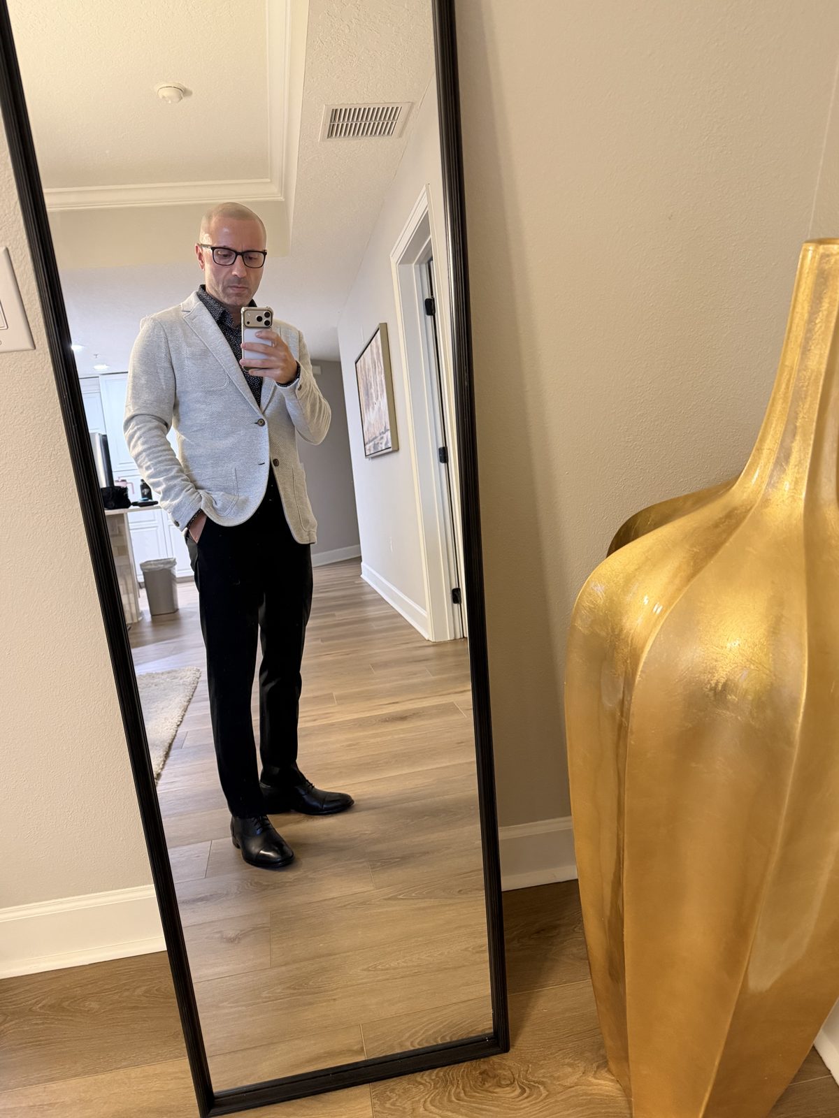 Feras in a light blazer and dark pants, standing in front of a hallway mirror, holding his phone in one hand