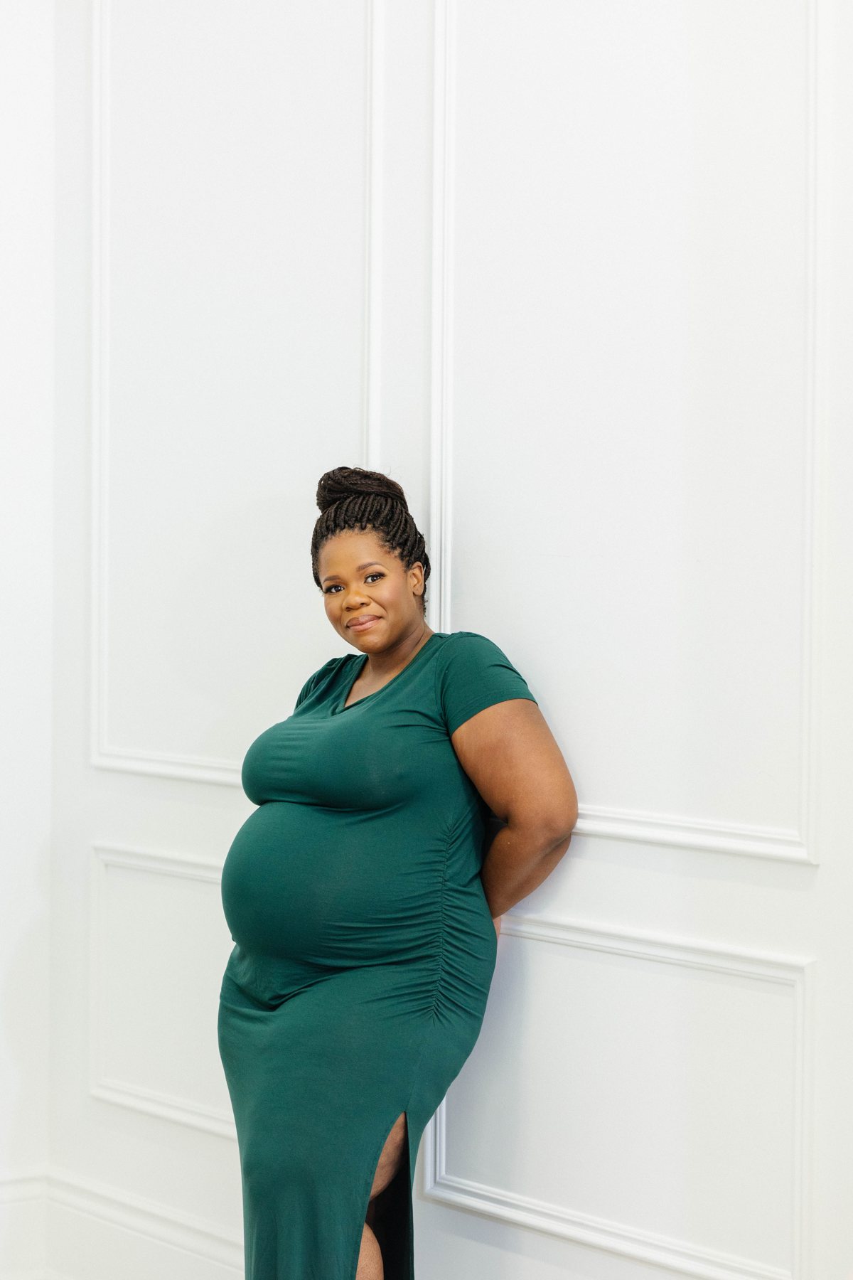 Sophie leaning against a white wall, pregnant, in a green dress