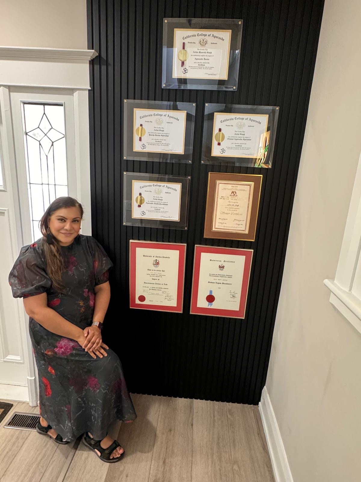 Dr. Arlini Singh standing beside her framed credentials: California College of Ayurveda, Matric, University of Durban-Westville, and University of Natal.