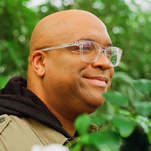 Mike Veny smiling outdoors in front of green foliage