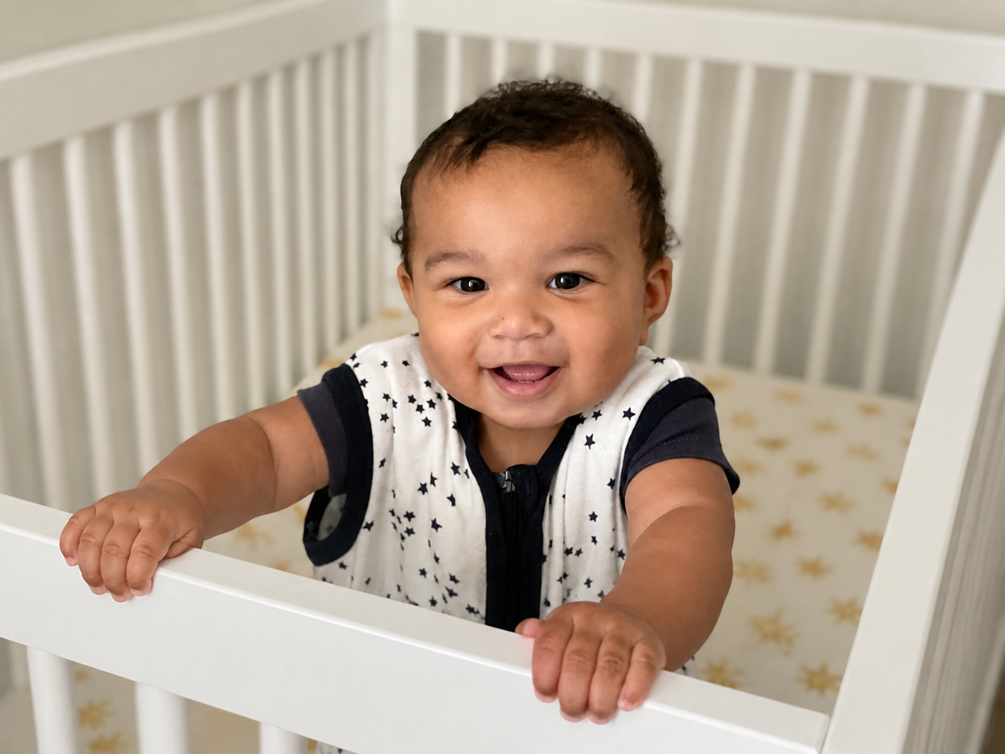 Smiling baby wearing a sleep sack while standing in a white crib