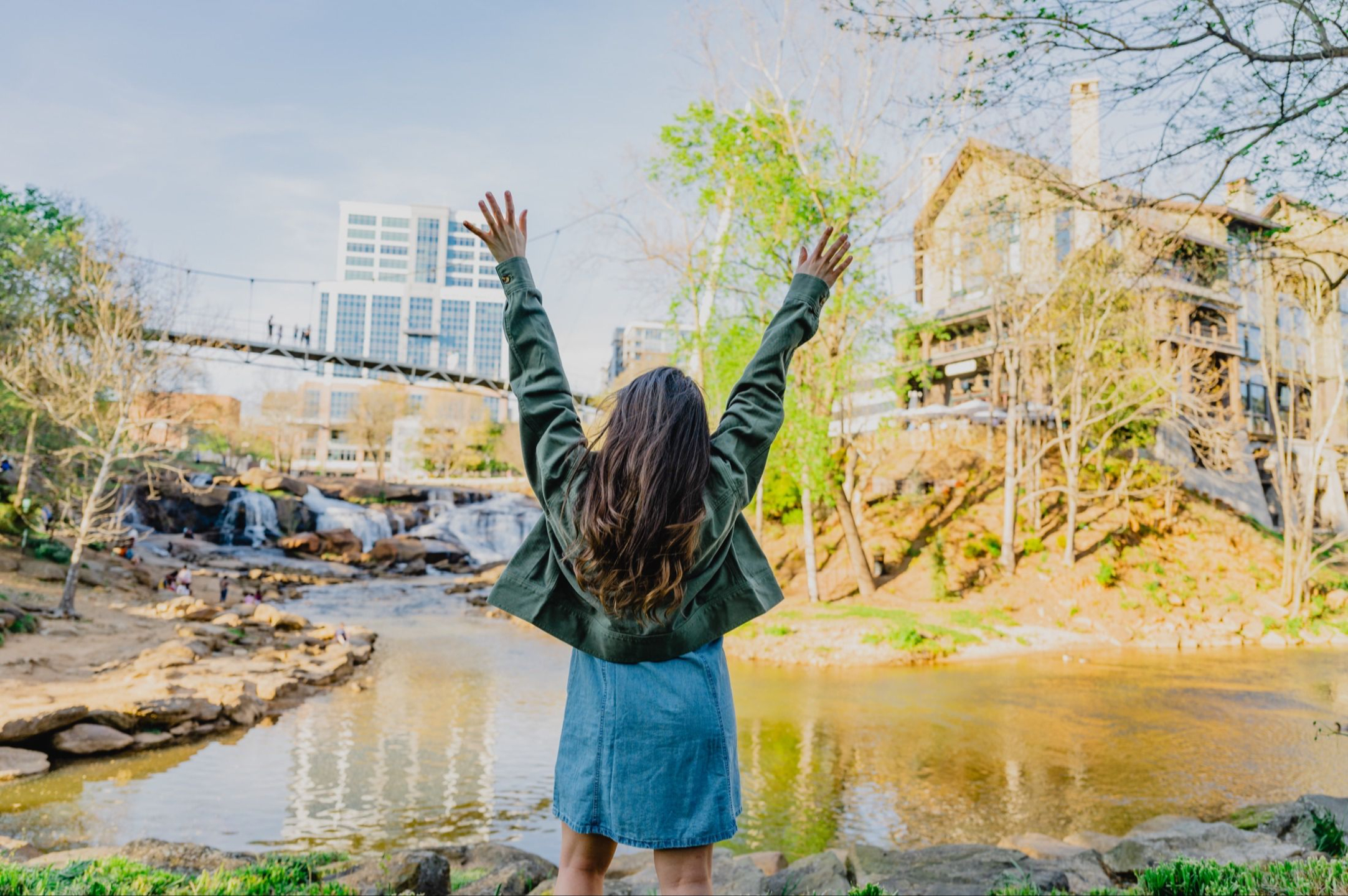 Jacqueline standing beside a river with her arms raised, facing a waterfall and footbridge