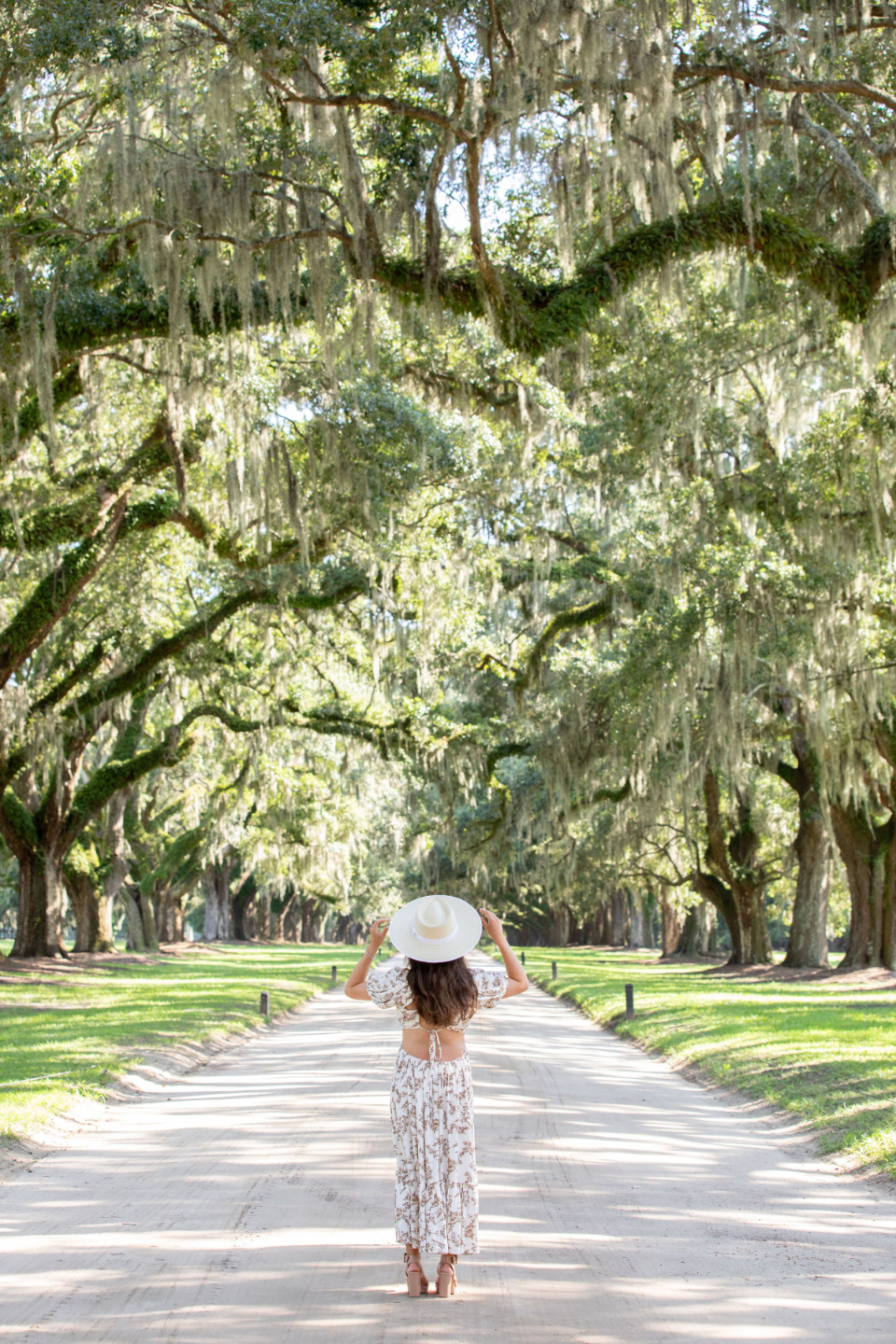 Jacqueline in a long floral dress standing on a dirt path beneath an avenue of live oaks draped in Spanish moss
