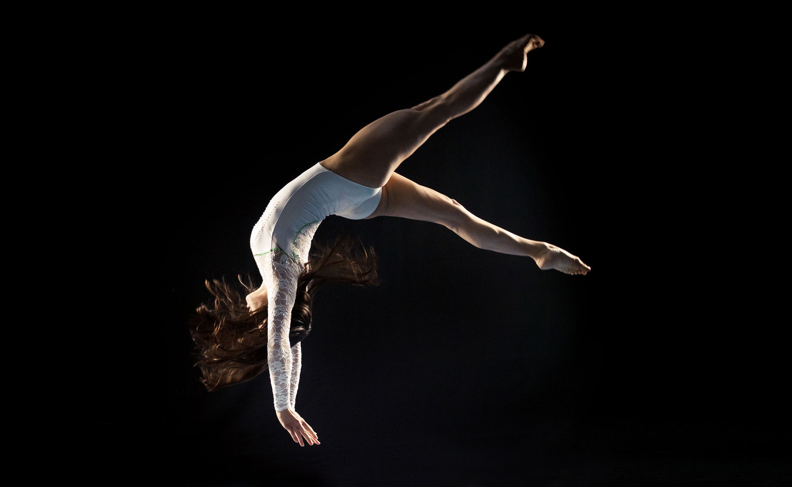 Gymnast in white leotard performs a backflip against a black background, hair flowing, conveying grace and dynamic motion.