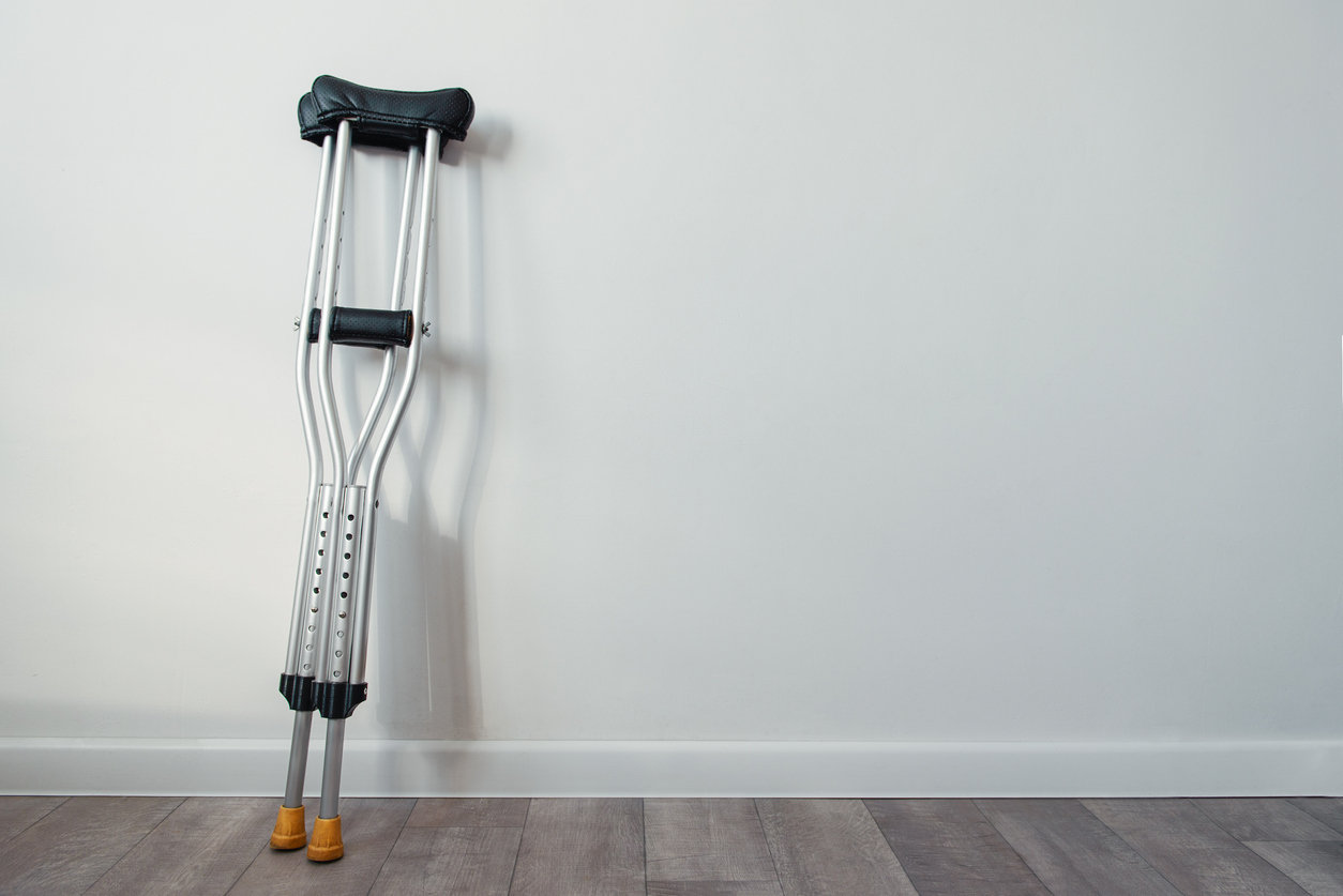 A single pair of crutches rests against a plain white wall on a wooden floor, evoking a serene, minimalist atmosphere.