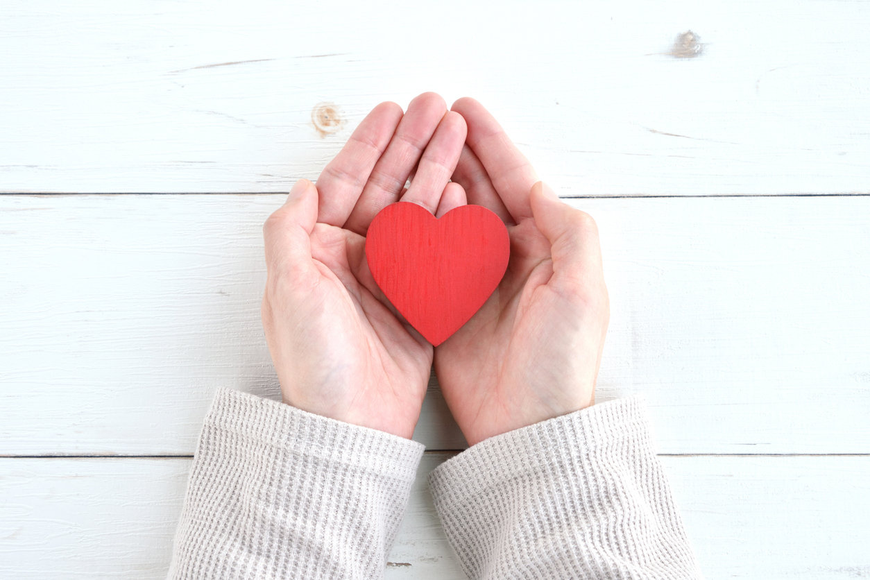 Hands in a white sweater hold a red heart-shaped object against a white wooden background, conveying a sense of care and warmth.