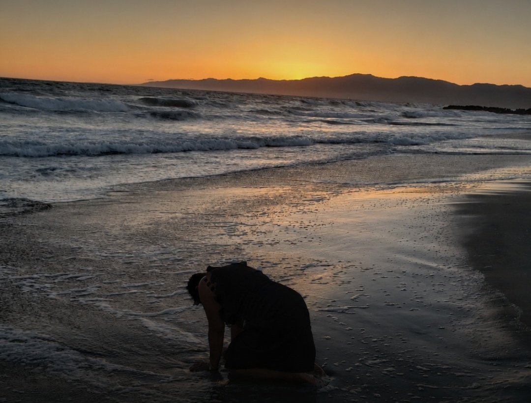 A woman bowed low in surrender at the shoreline at sunset